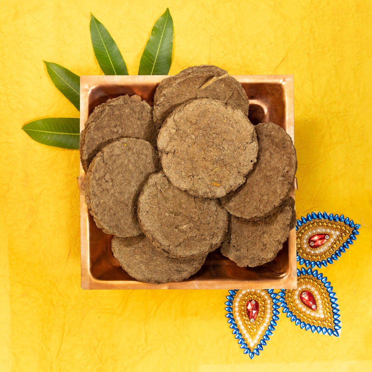 A top-down view of dark brown round herbal cakes, likely amla or amla powder cakes, arranged in a small square wooden dish on a textured yellow background with decorative green leaves and embroidered motifs.
