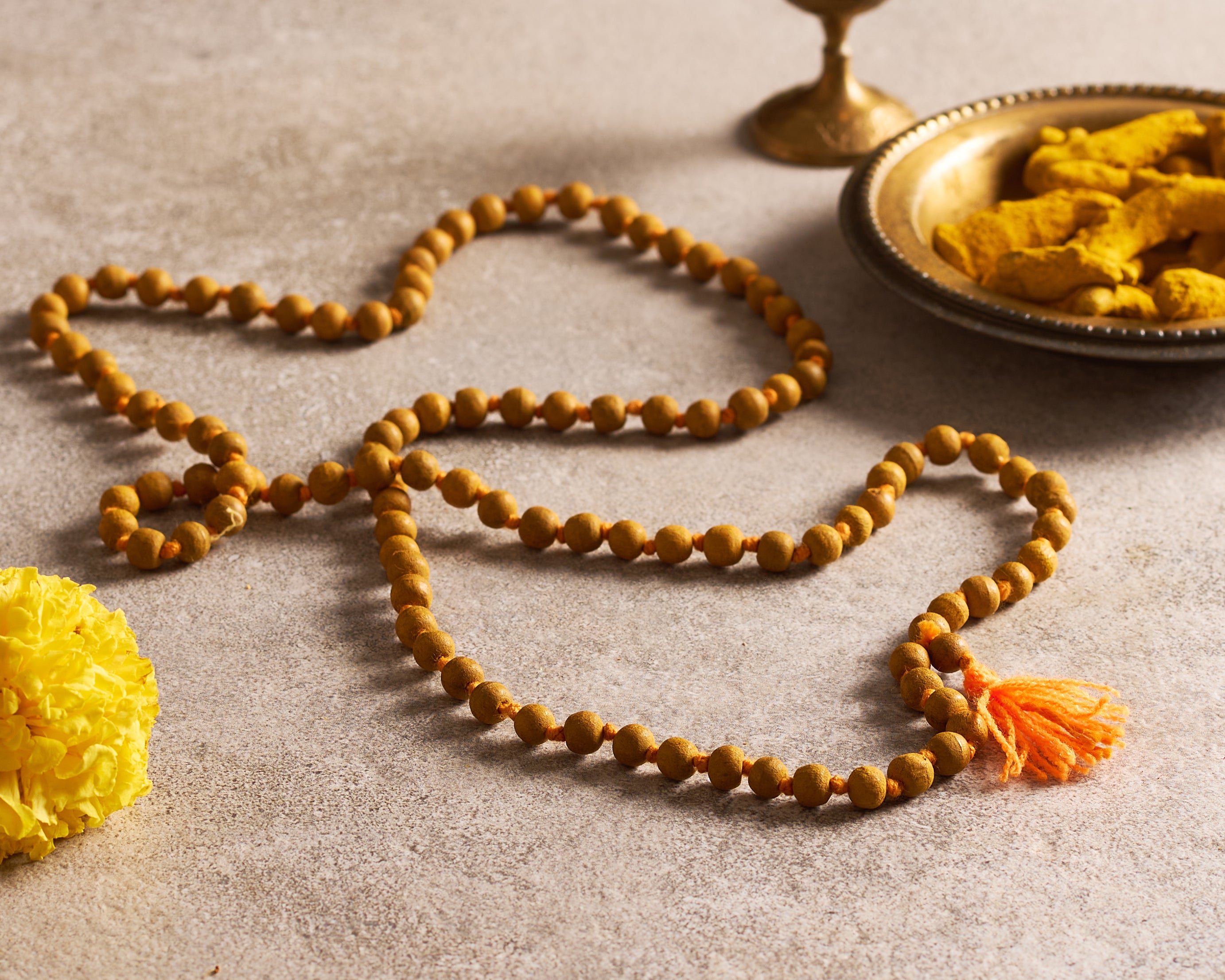 Brown beaded necklace with an orange tassel on a beige surface, accompanied by yellow flowers and a small dish.