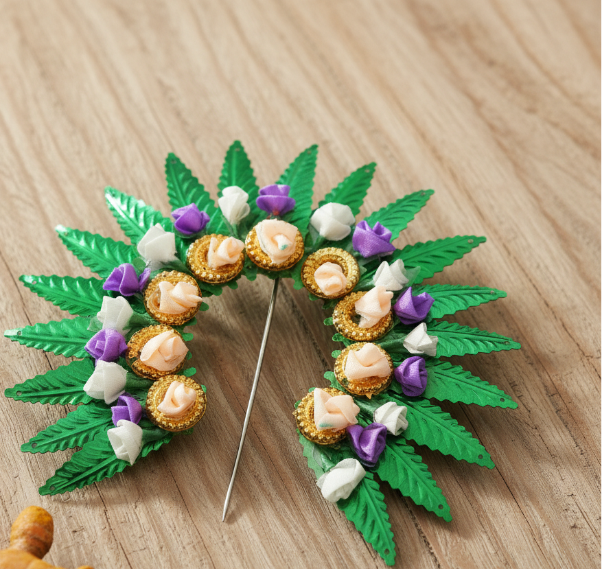 A decorative brooch or pin shaped like a leafy wreath, featuring metallic green leaves, peach and white paper flowers, and purple accents, with a silver pin stem in the center, shown on a dark gray gradient background.