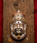 A silver-toned metal wall plaque depicting a Hindu deity's face, adorned with colorful gemstone-like accents and a crown, mounted on a rustic wooden board with red fabric borders.