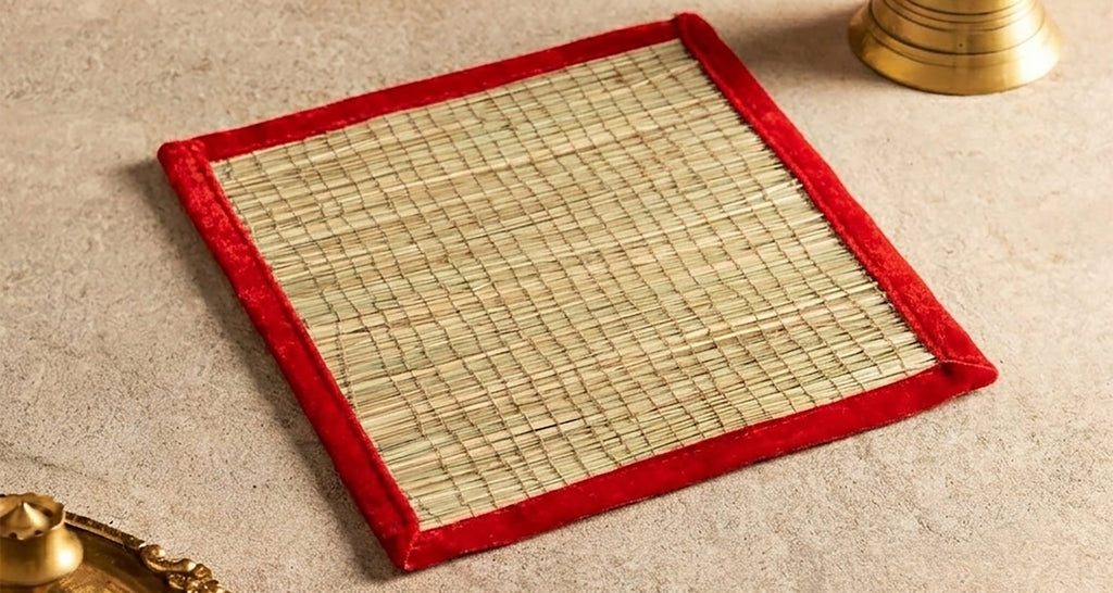 A square woven mat with a red velvet border, styled on a beige textured surface with traditional brass props and marigold flowers in the background.