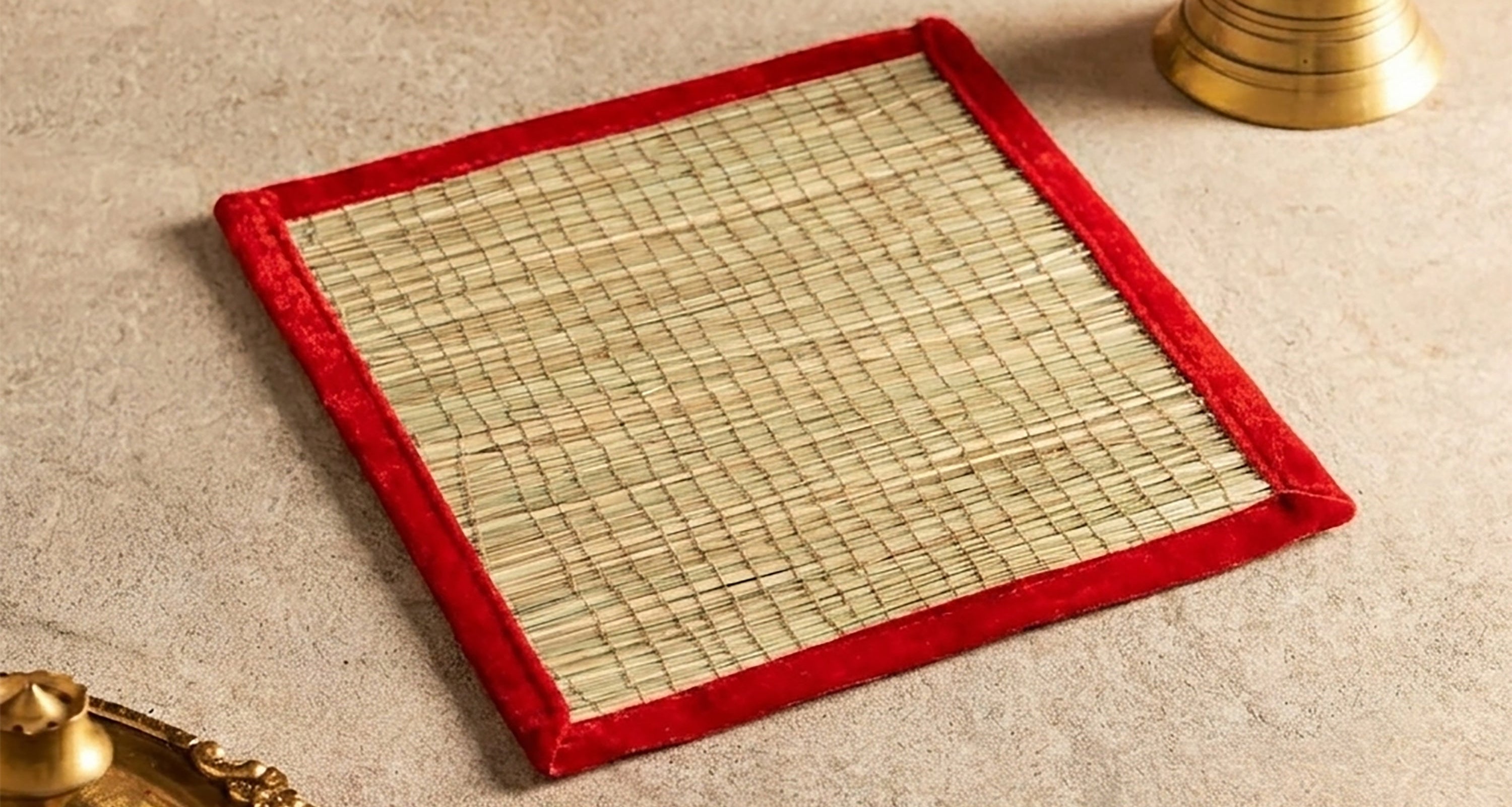 A square woven mat with a red velvet border, styled on a beige textured surface with traditional brass props and marigold flowers in the background.