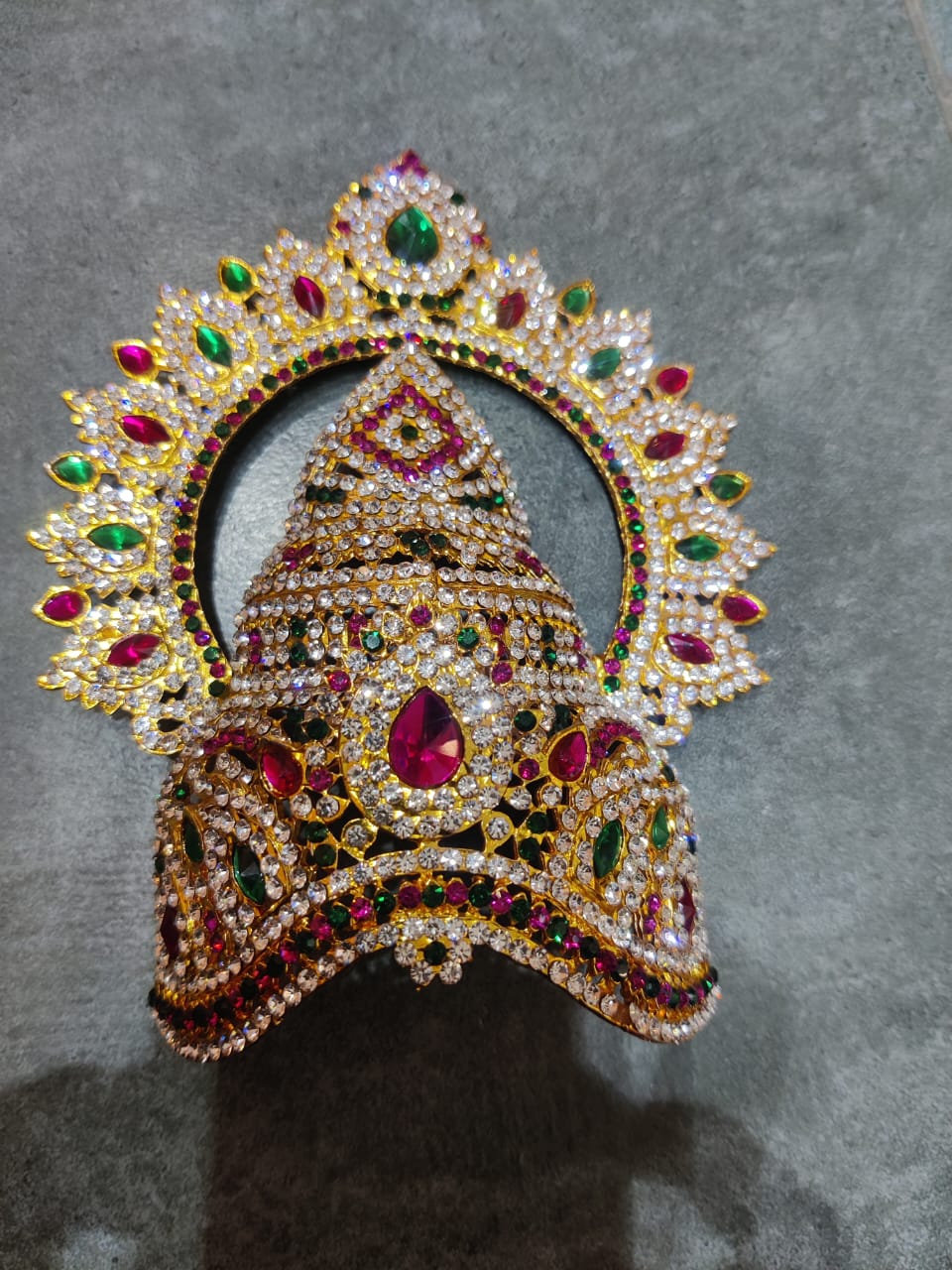 A multicolored stone keerita with arch design, featuring metal casing and adorned with various colored stones, intended for decorating a god.