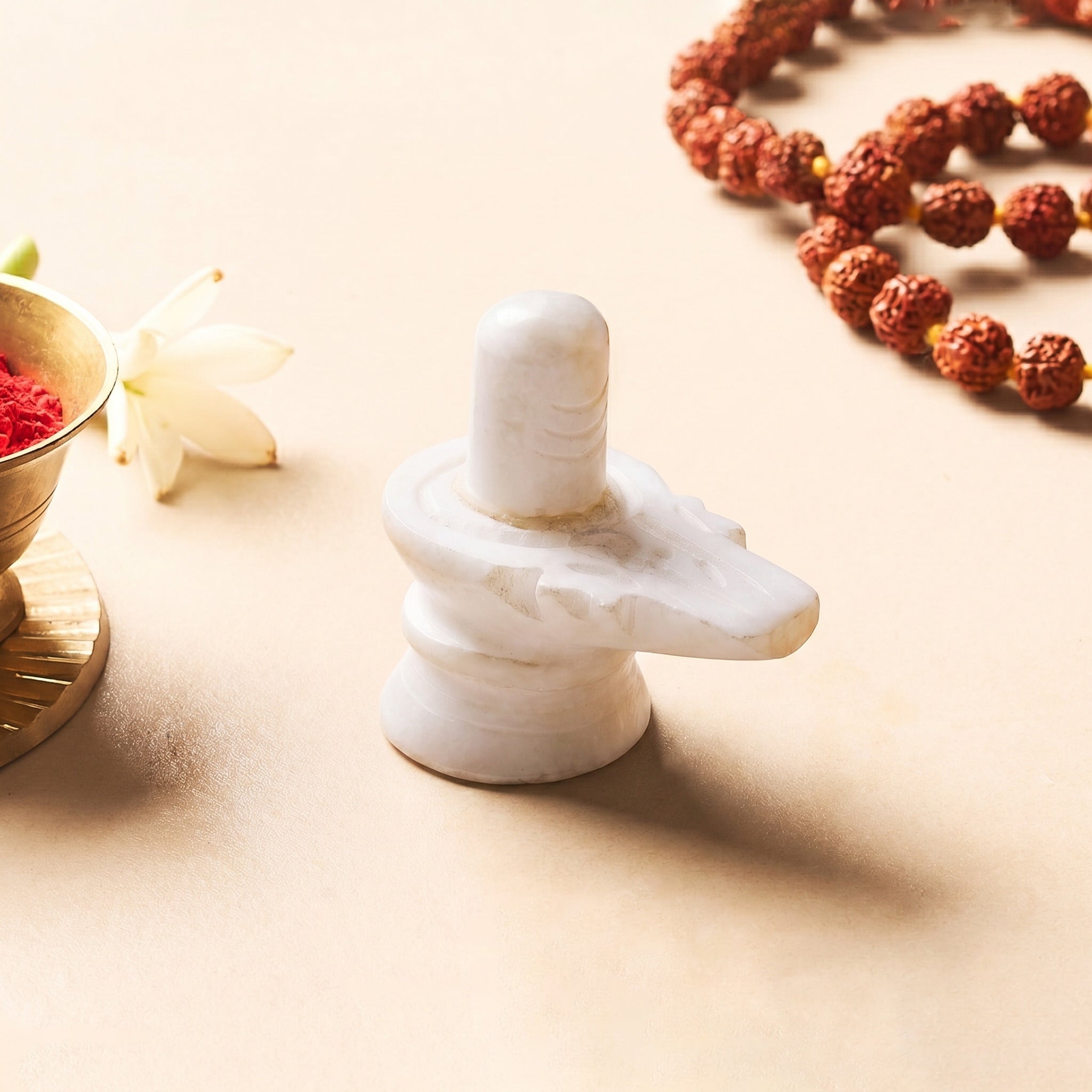 White marble Shiva lingam on a beige surface with red and white flowers and a rosary in the background.
