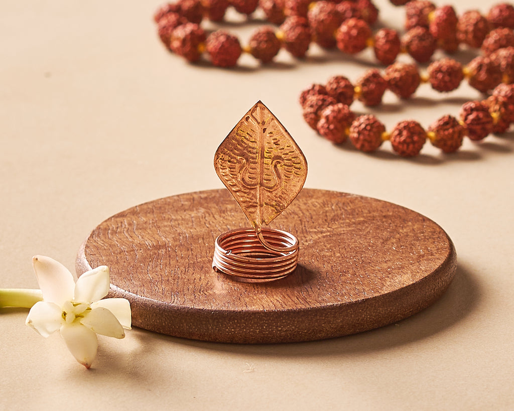 Decorative copper leaf and ring on a wooden stand with rudraksha beads in the background.