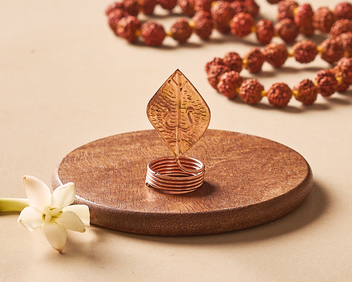 Decorative copper leaf and ring on a wooden stand with rudraksha beads in the background.