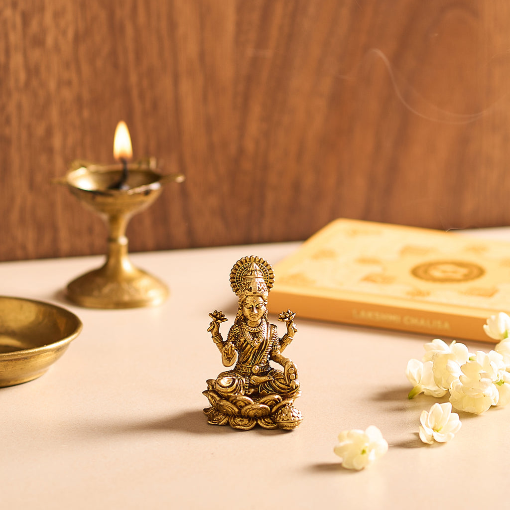 A prayer altar with an altar book, fresh white jasmine flowers and a brass lamp next to an antique golden brass Lakshmi idol on a lotus throne.