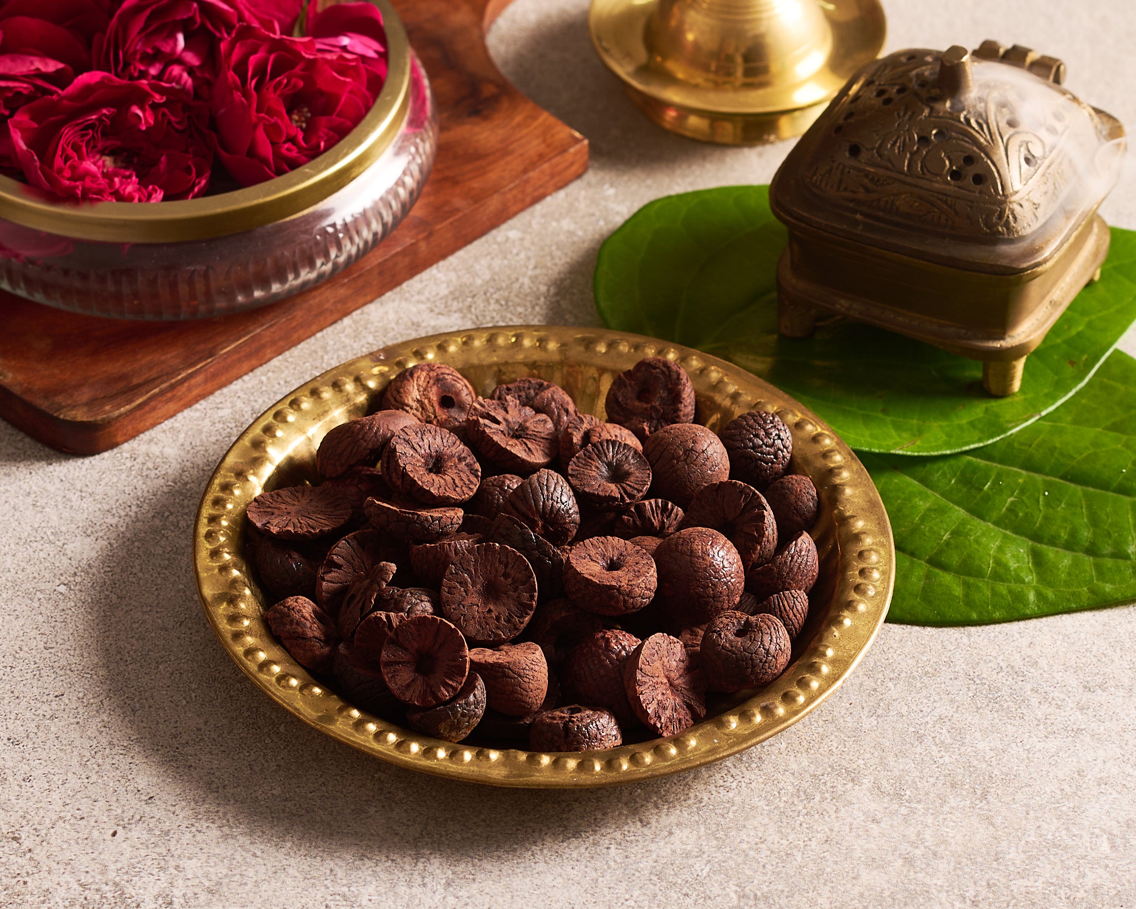 Brass bowl with dried fruits on a light surface with green leaves and a decorative item.