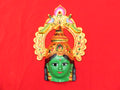 A decorative aluminum item featuring the face of Ashta Lakshmi, the Hindu goddess of wealth, with a vibrant green complexion and gold details against a red background.